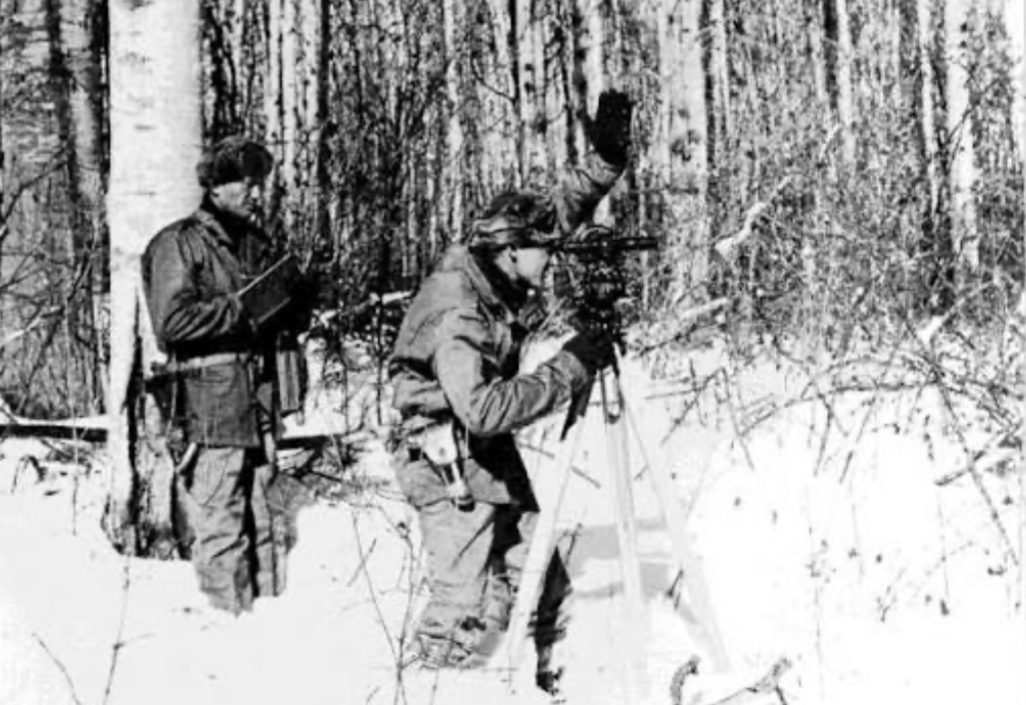 Unverified. A black-and-white photograph of two soldiers in winter clothing standing in deep snow in a birch forest. One holds a notebook. The other bends to a surveyor's instrument on a tripod and raises a gloved hand as a signal.